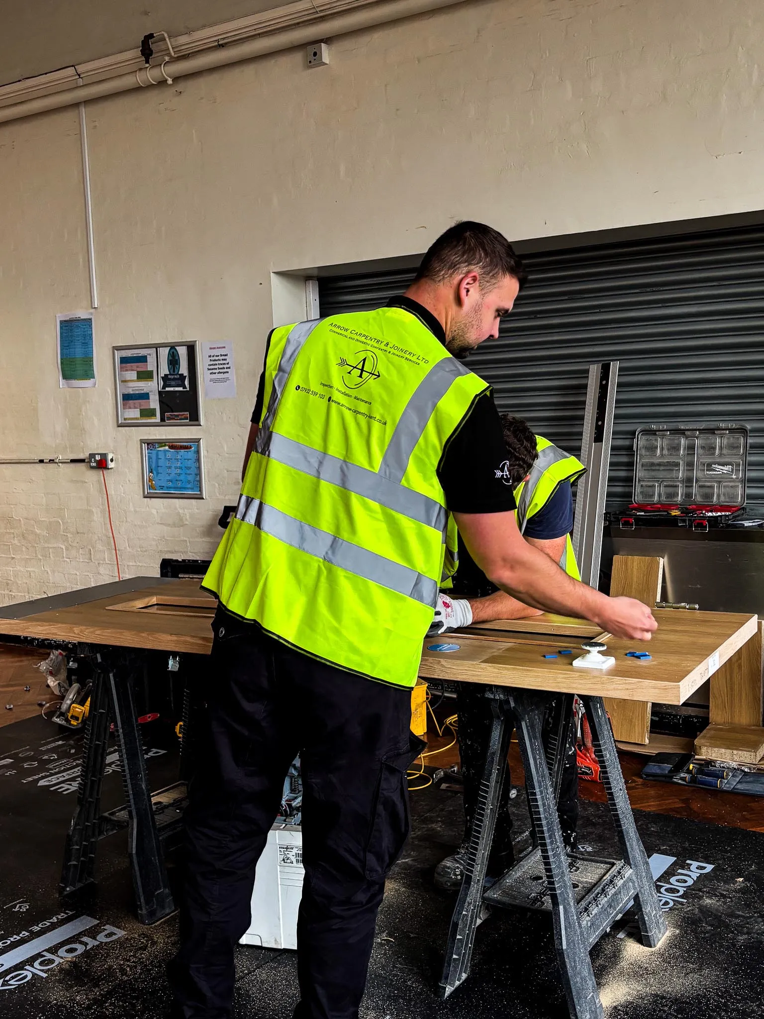 Two men in high-visibility vests working on a wooden door in a workshop setting.