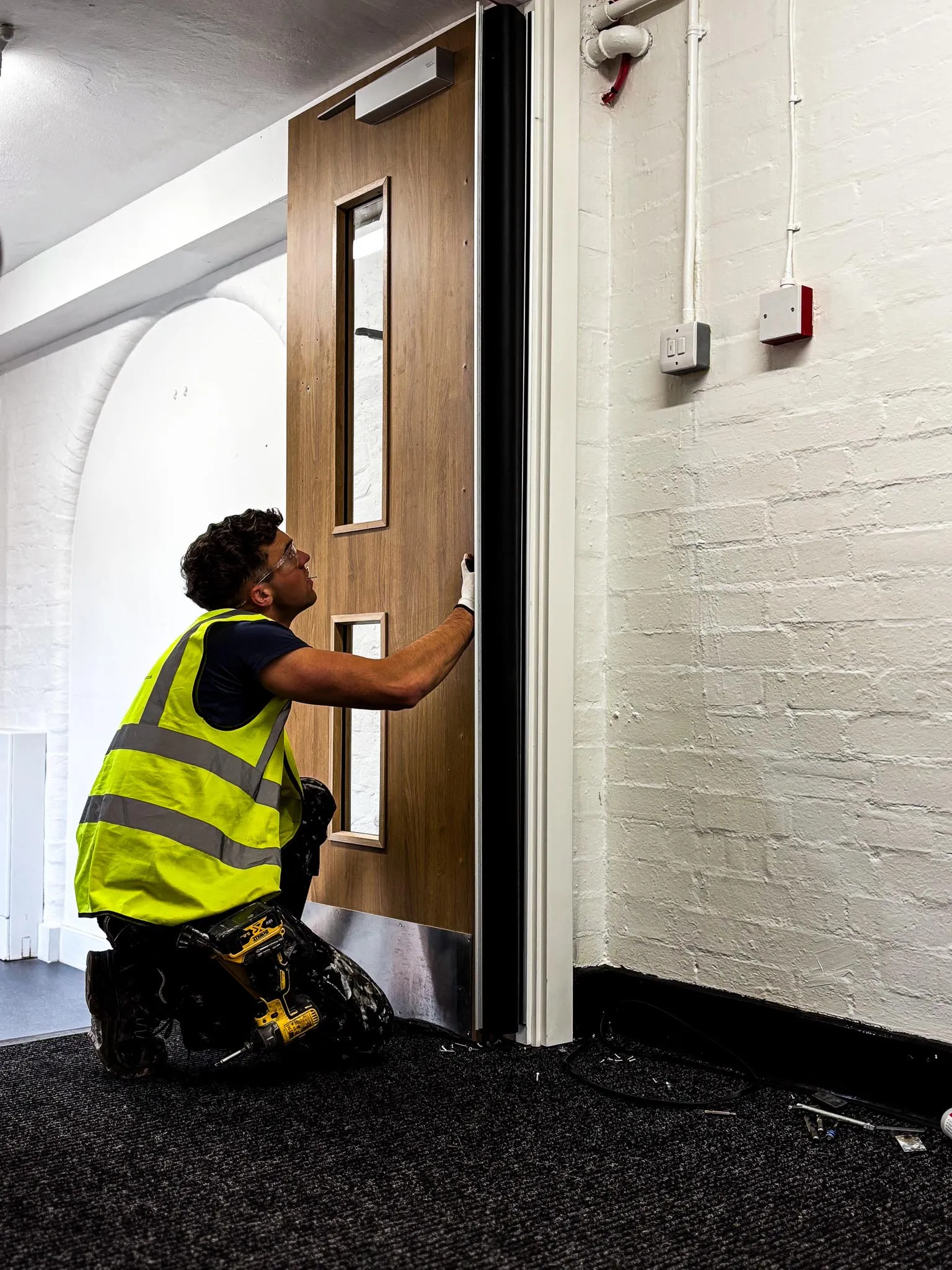 Construction worker in a yellow safety vest installing or adjusting a wooden door inside a building.