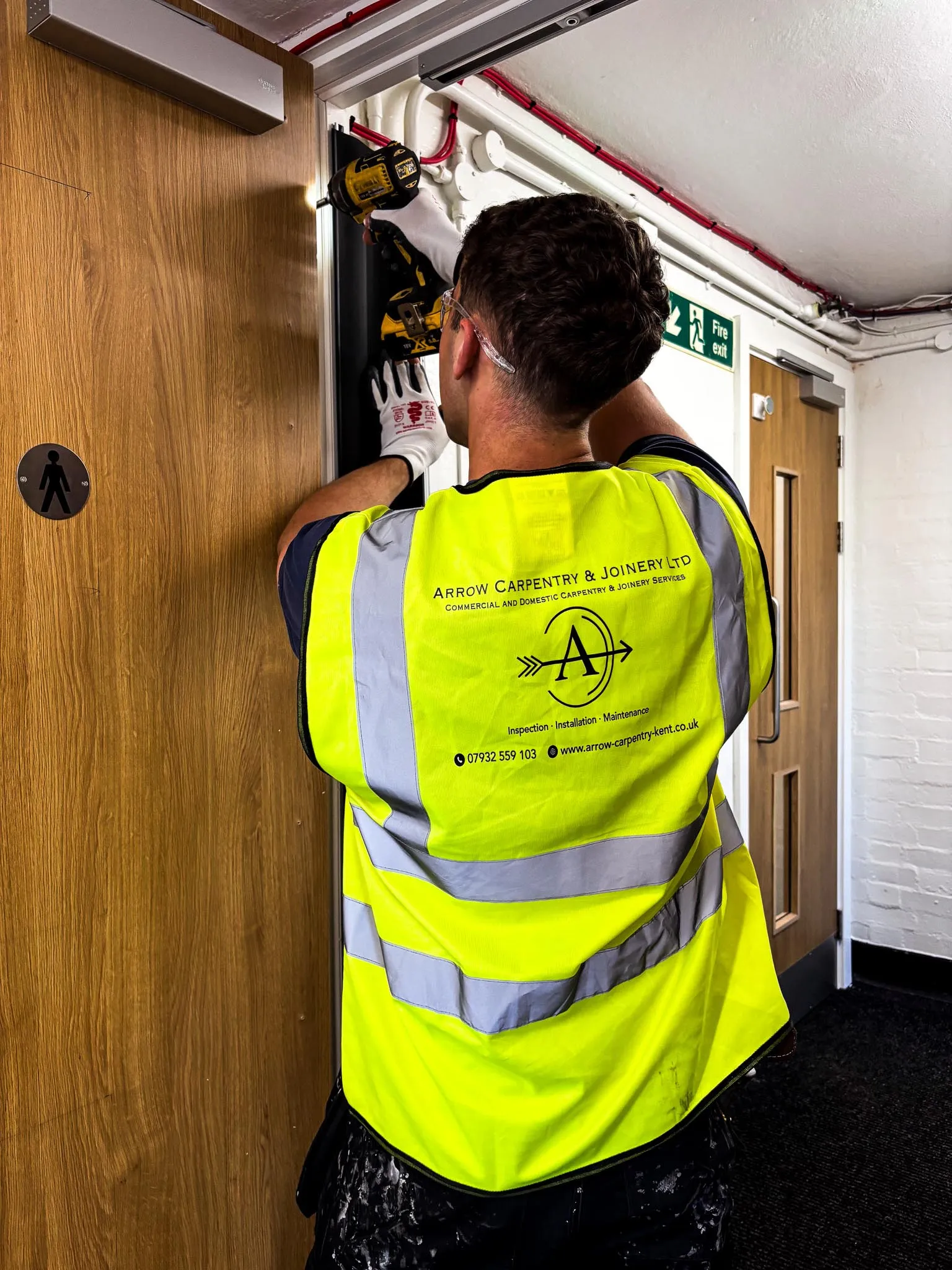 Carpenter wearing a high-visibility vest and safety glasses uses a drill to install a door frame in an indoor hallway.