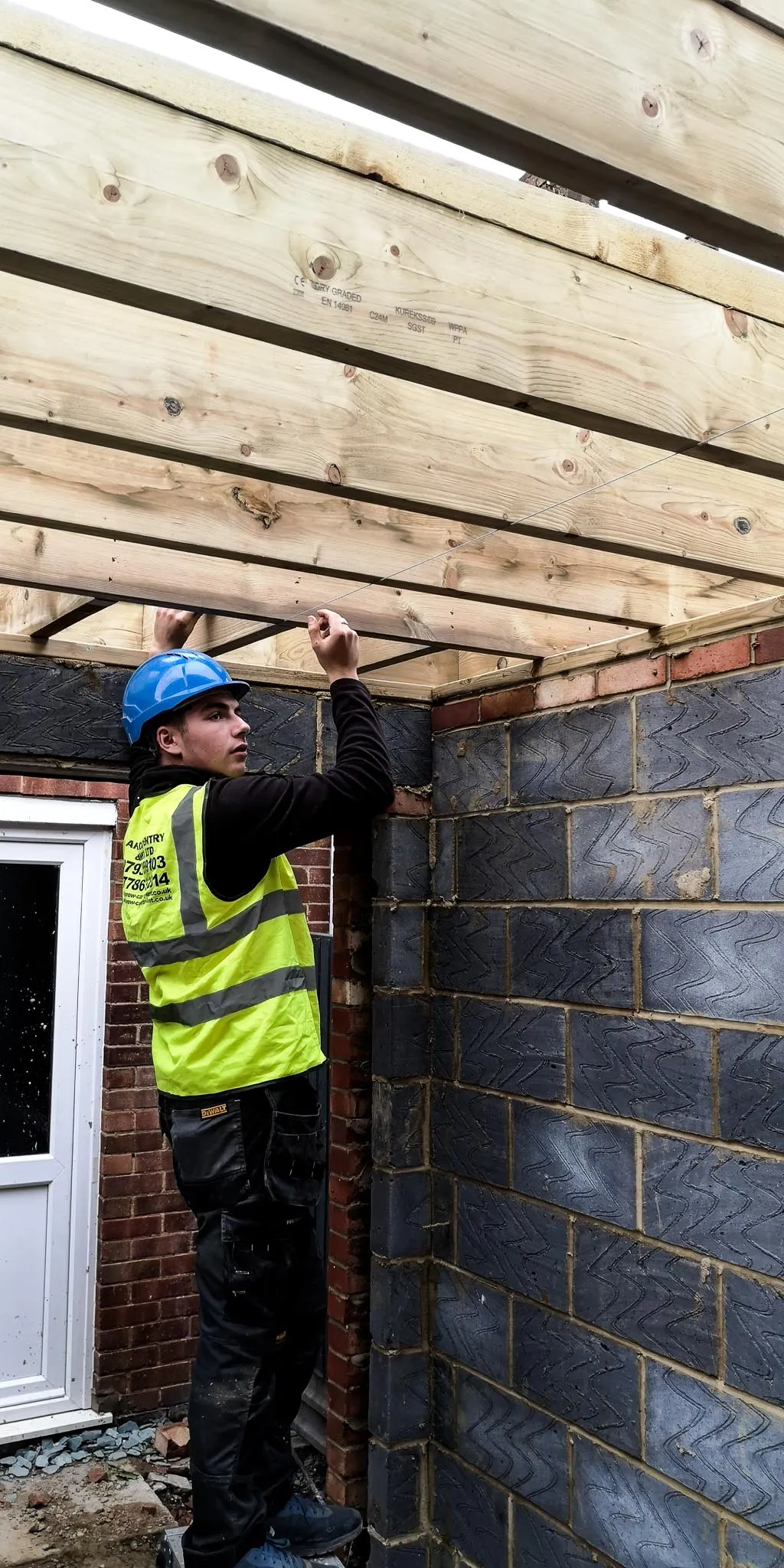Construction worker in a blue hard hat and yellow safety vest working on wooden beams inside a partially built brick and cinder block structure.