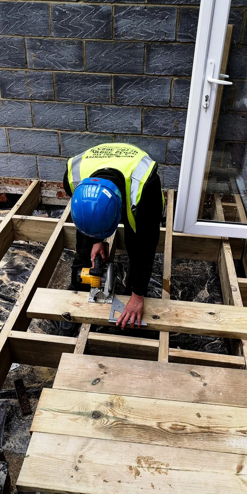 Construction worker wearing a blue helmet and yellow safety vest cutting a wooden plank with a circular saw while using a carpenter's square.