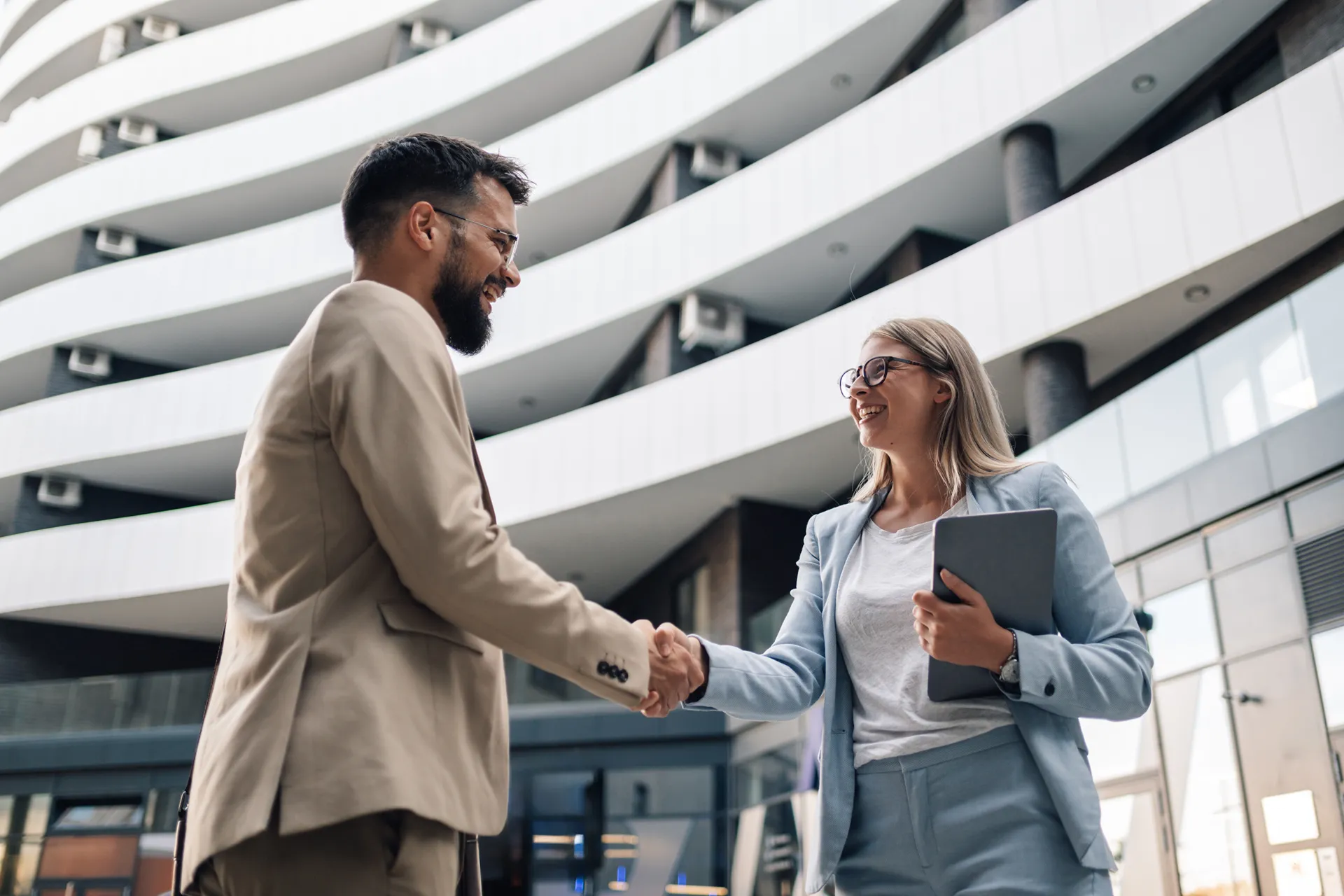 Smiling man and woman in business attire shaking hands outside a modern office building.