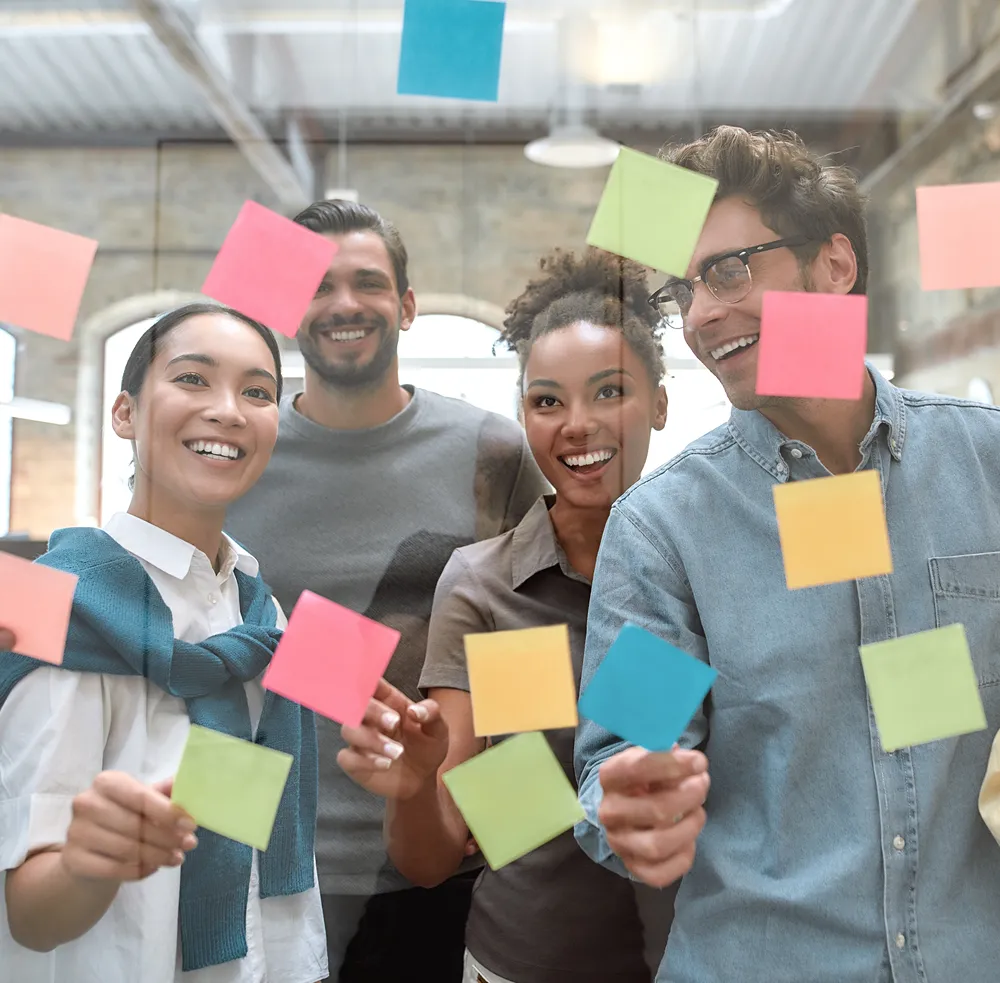 Diverse group of four young professionals smiling and placing colorful sticky notes on a glass wall in a modern office.