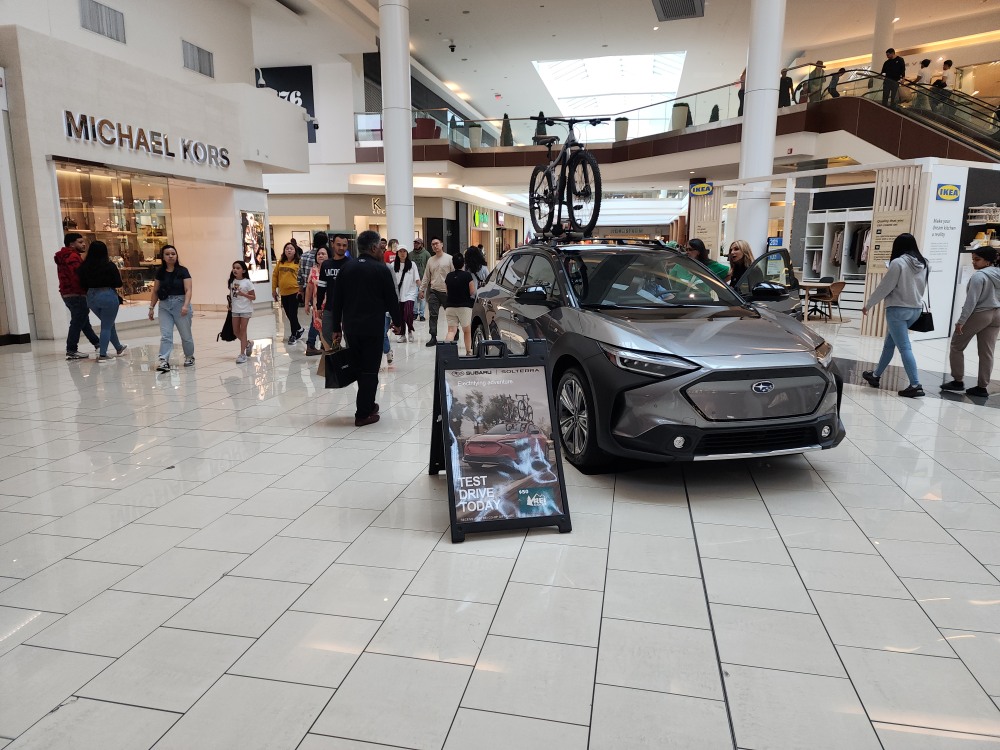 Silver Subaru Solterra electric vehicle on display in a shopping mall with a bike mounted on its roof and a sign promoting test drives.