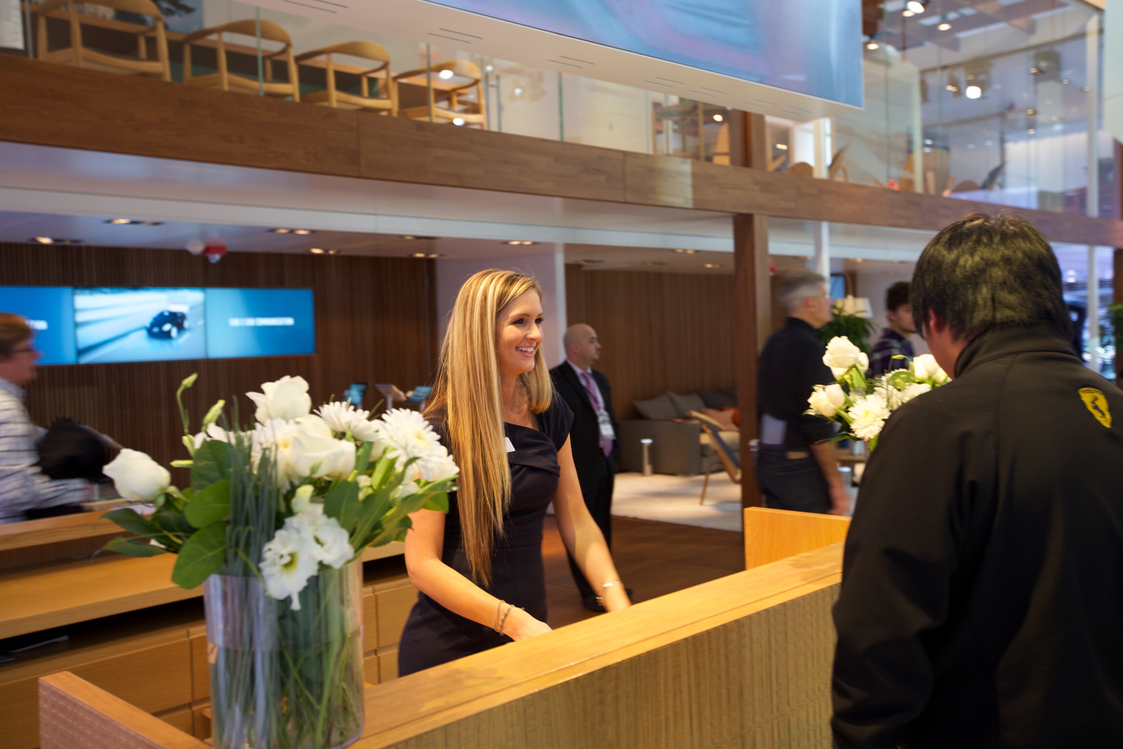 Smiling woman standing behind a wooden reception desk with a vase of white flowers, greeting a man in a black jacket.