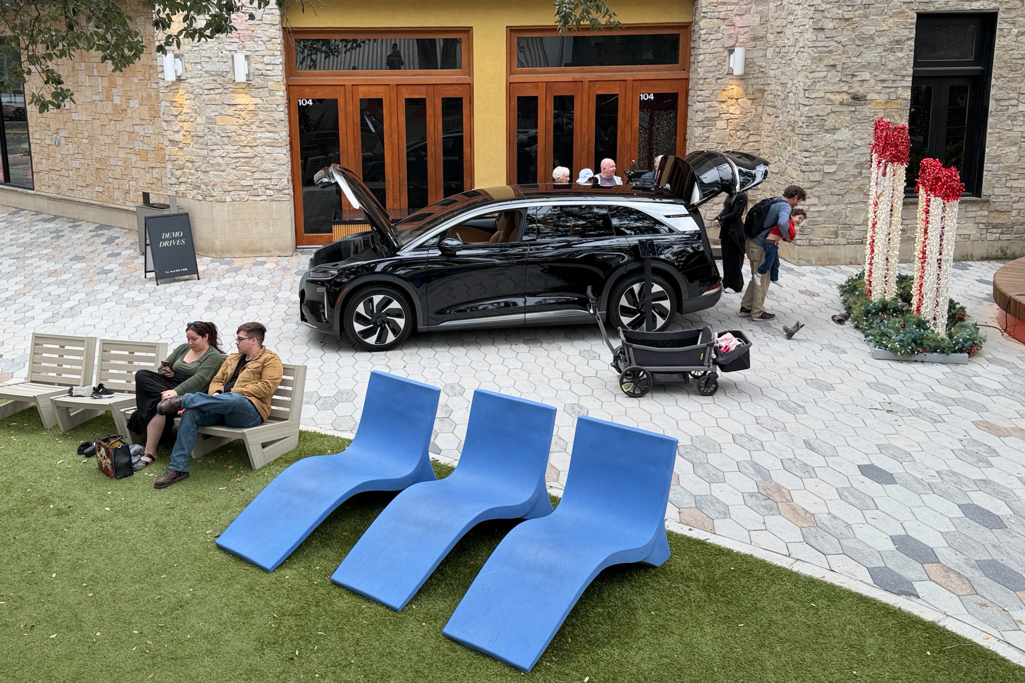 Black electric SUV with open hood and trunk parked on a hexagonal tiled plaza near a building entrance, a man holding a child nearby, two people sitting on a bench on grass, and three blue lounge chairs on the grass.