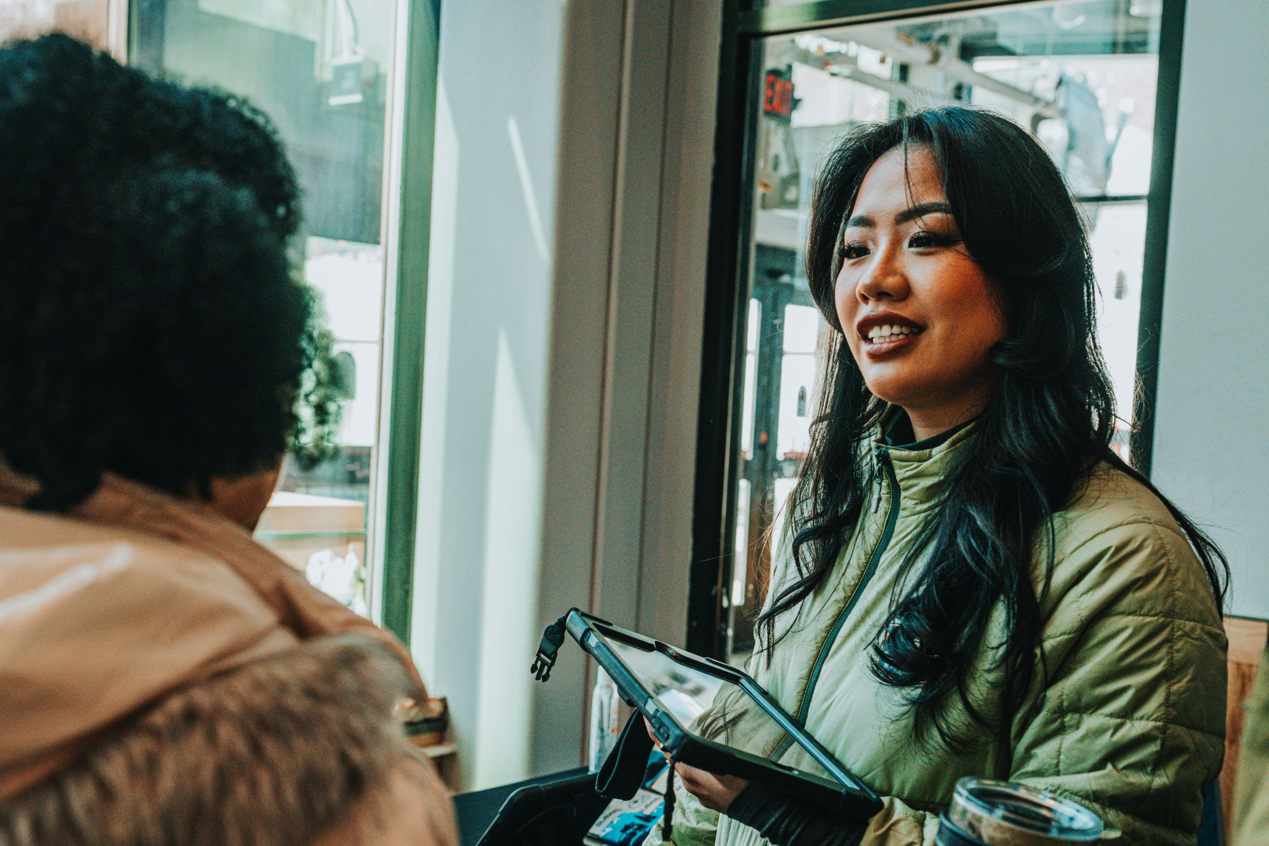 Smiling woman in a green jacket holding a tablet and talking to another person indoors near a window.