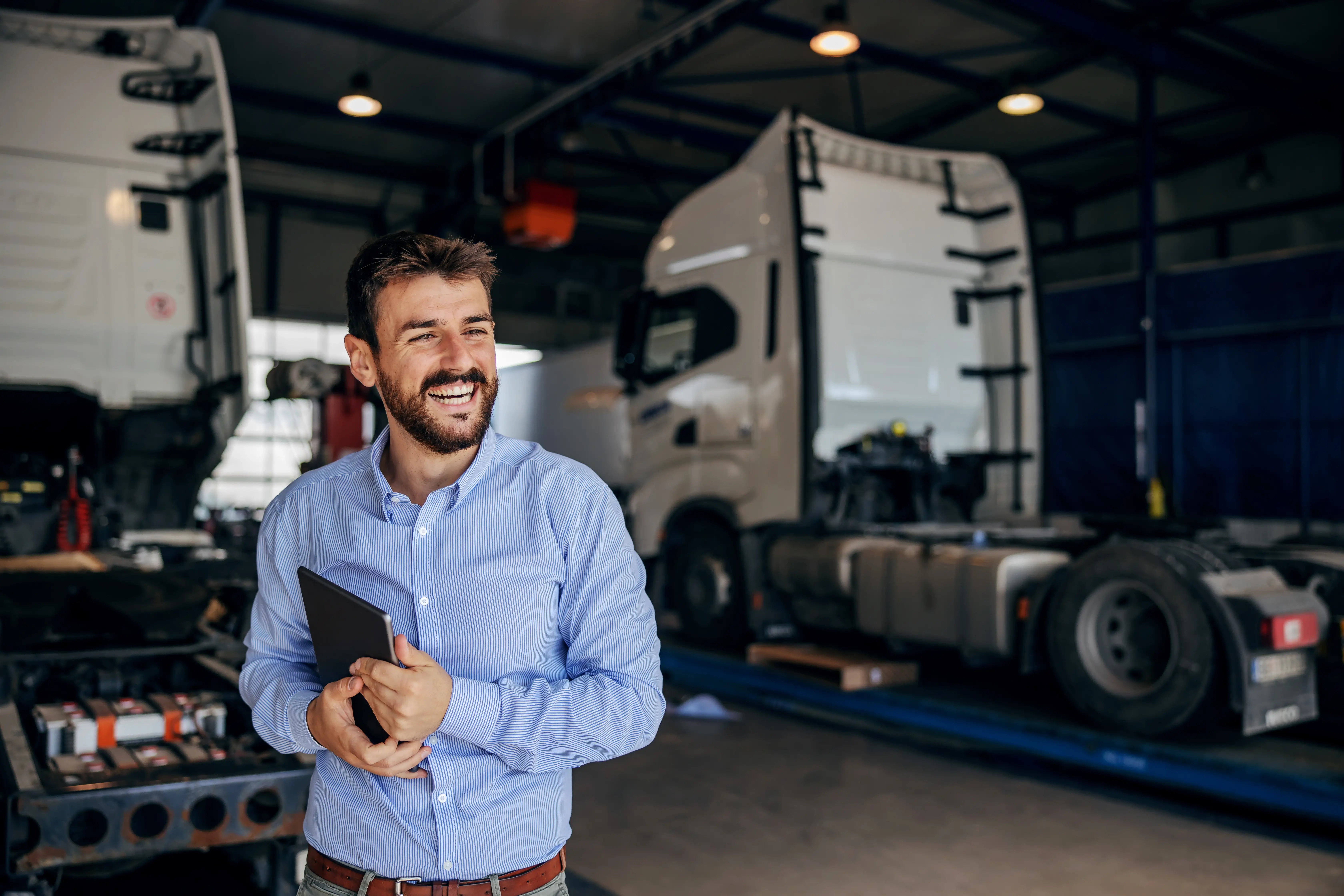 Smiling man in a blue striped shirt holding a tablet inside a truck repair garage with large trucks in the background.