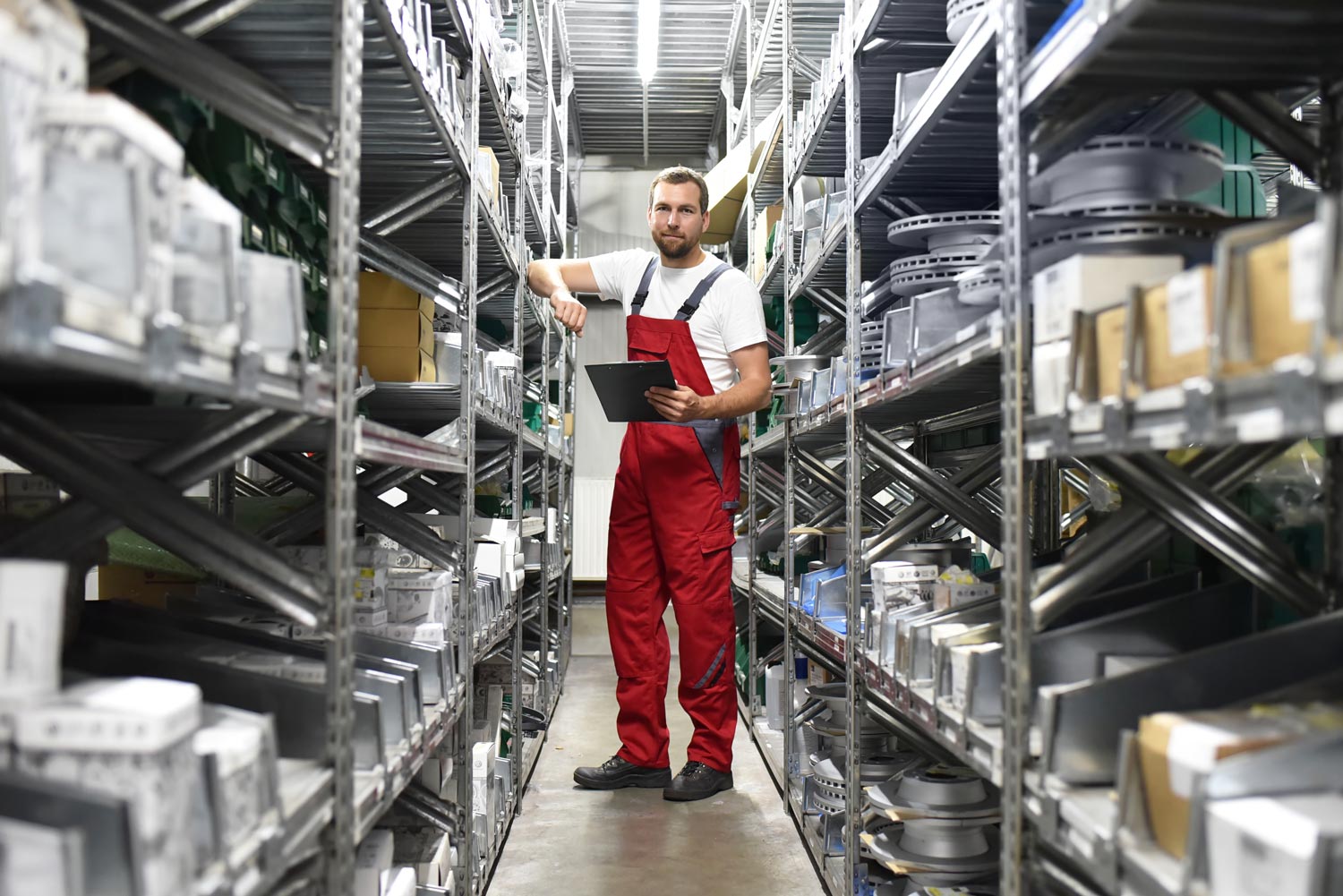Warehouse worker in red overalls holding clipboard and checking inventory between metal shelves filled with parts.