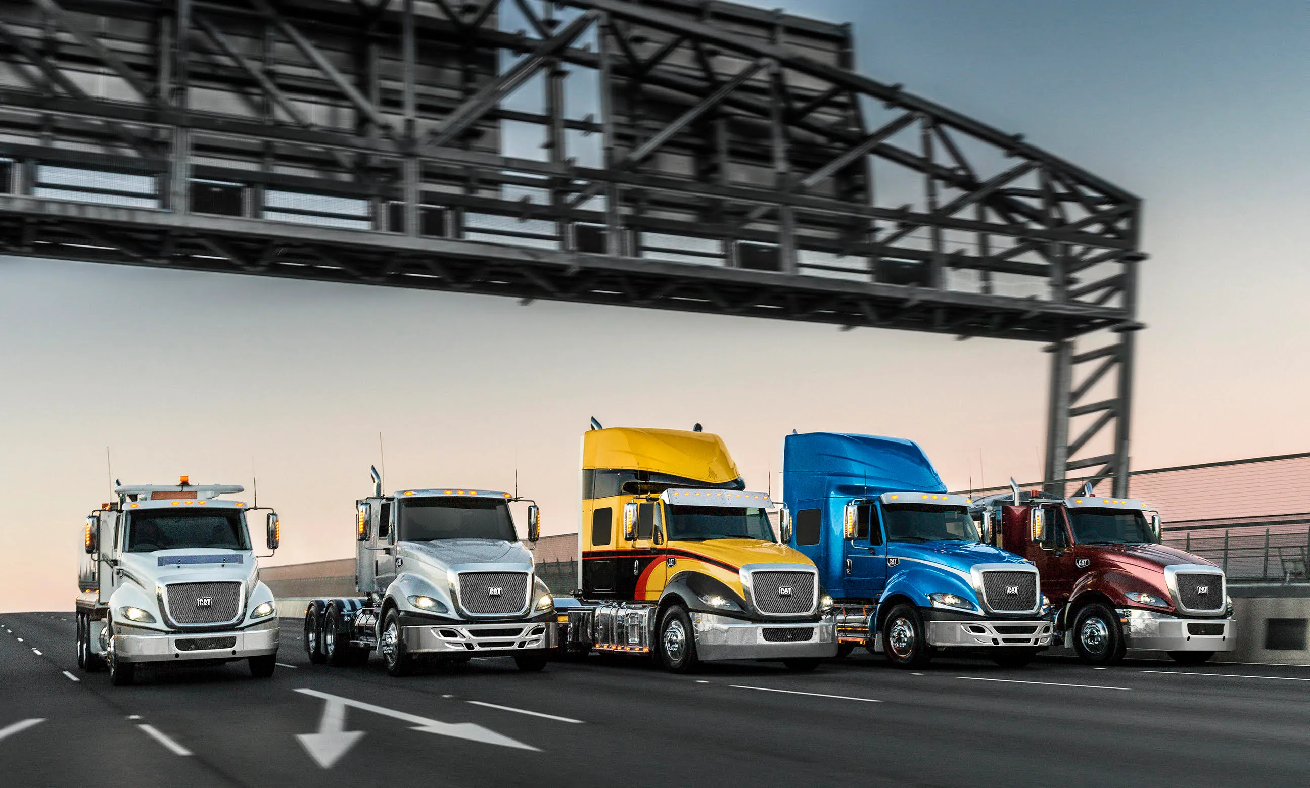 Five CAT semi trucks in silver, yellow, blue, and red lined up on a highway under a metal overhead structure at dusk.