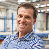 Smiling man wearing a checked shirt in an industrial or warehouse setting.