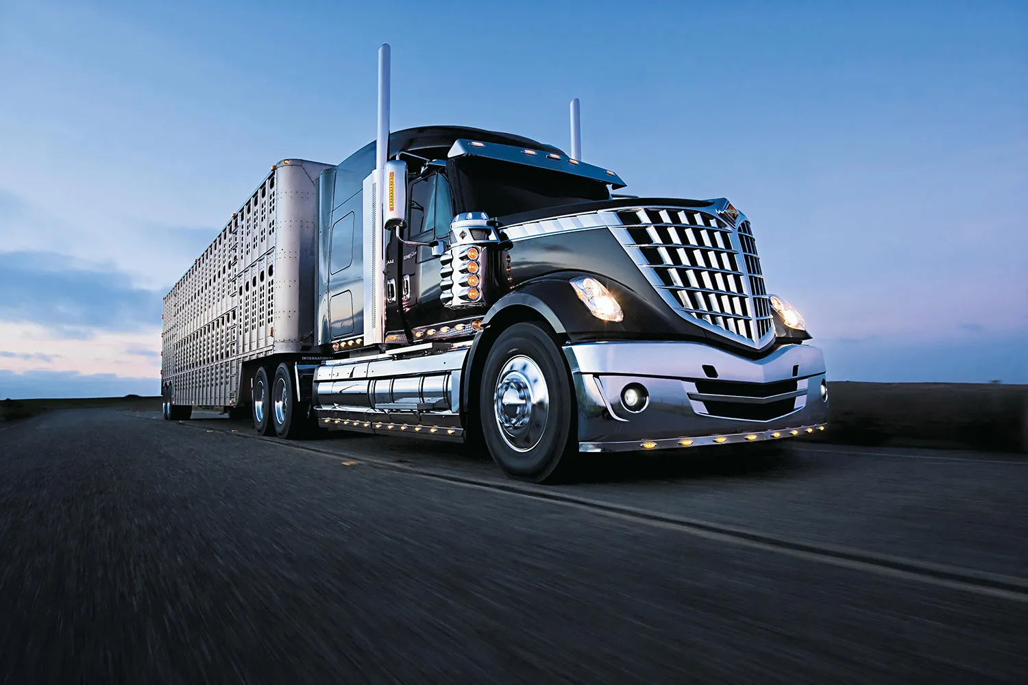 Black and chrome semi-truck with a livestock trailer driving on a road at dusk.