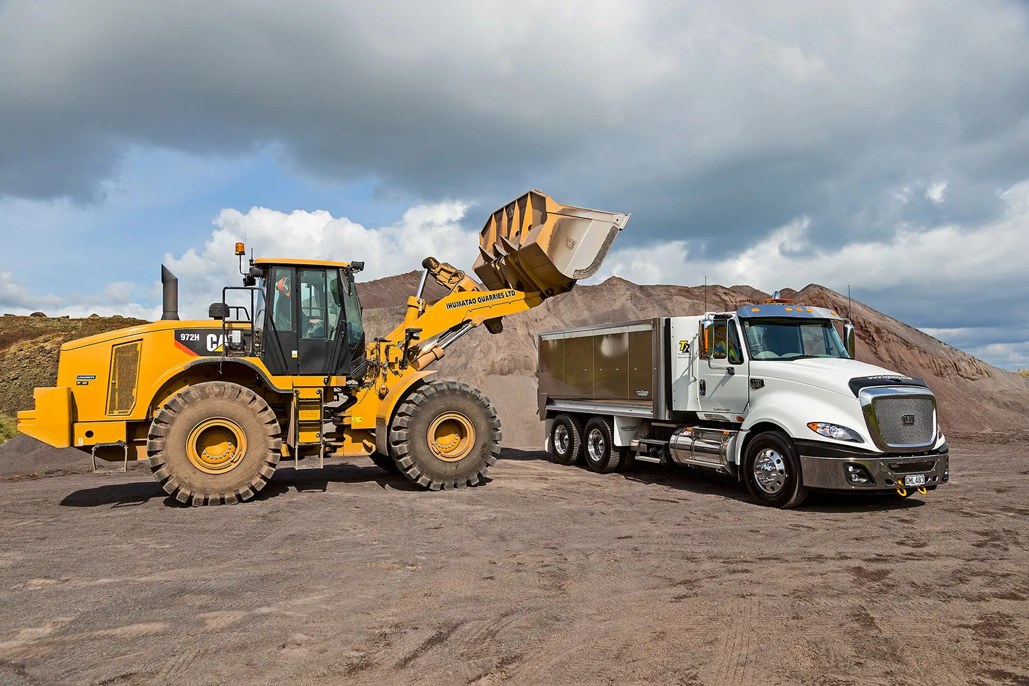 Yellow CAT 972H wheel loader loading gravel into a white dump truck at a quarry site with dirt piles and cloudy sky in the background.