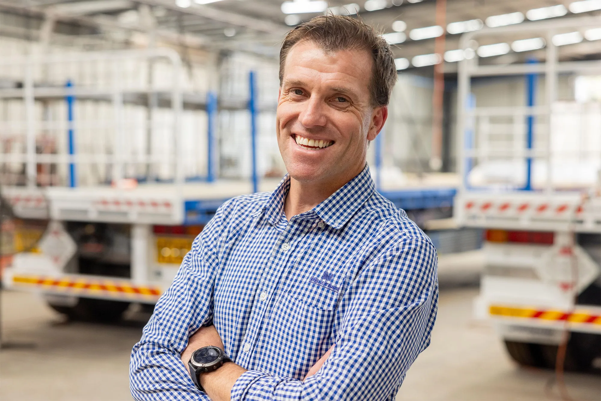 Smiling man in a blue checkered shirt with arms crossed standing in a warehouse with trucks in the background.
