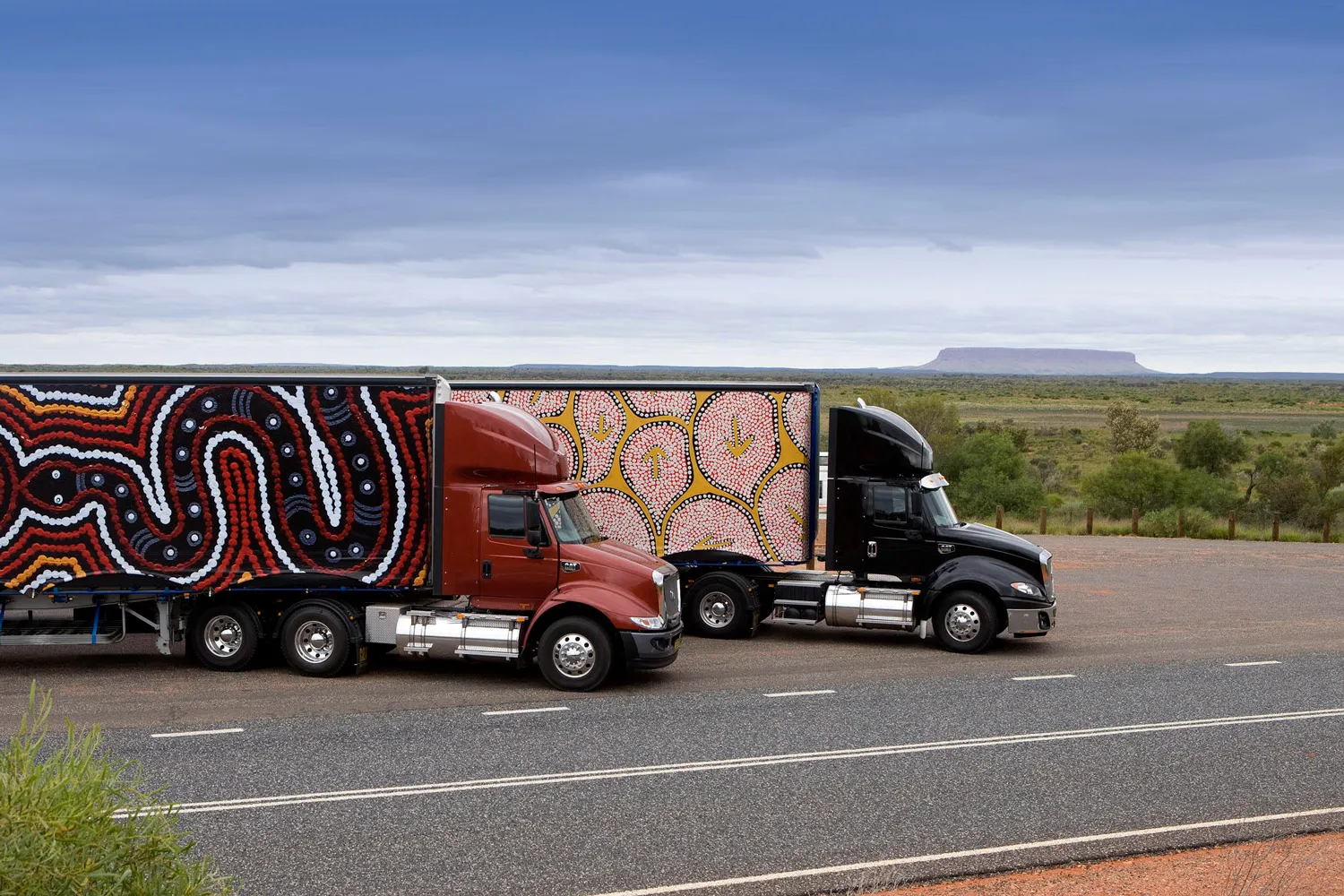 Two semi-trucks parked side by side on the roadside with trailers featuring vibrant Aboriginal dot paintings.