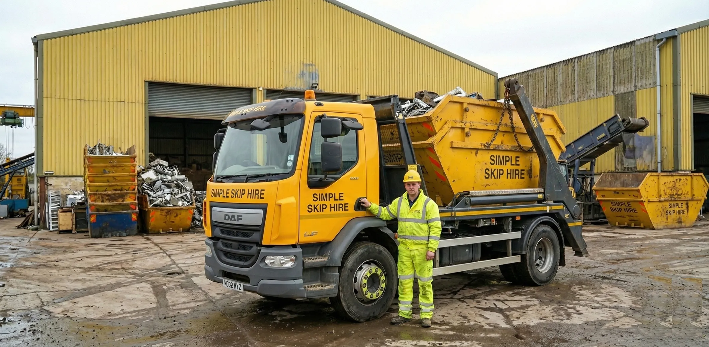 Worker in high-visibility clothing and helmet standing beside a yellow Simple Skip Hire truck with a skip container, at an industrial yard with yellow buildings and stacked metal scrap.