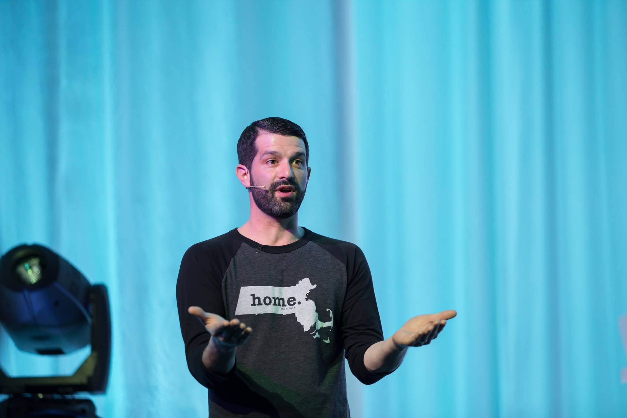 ZG Group - Zack delivering a keynote conference message. Man with beard speaking on stage wearing a black and gray shirt with a Massachusetts state outline and the word 'home.'