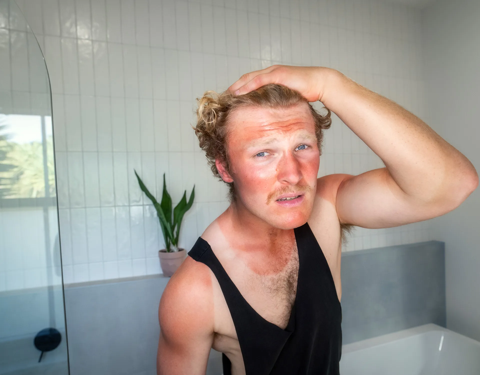 Man with sunburned face and shoulders wearing a black sleeveless shirt, holding his head with a concerned expression in a bathroom.