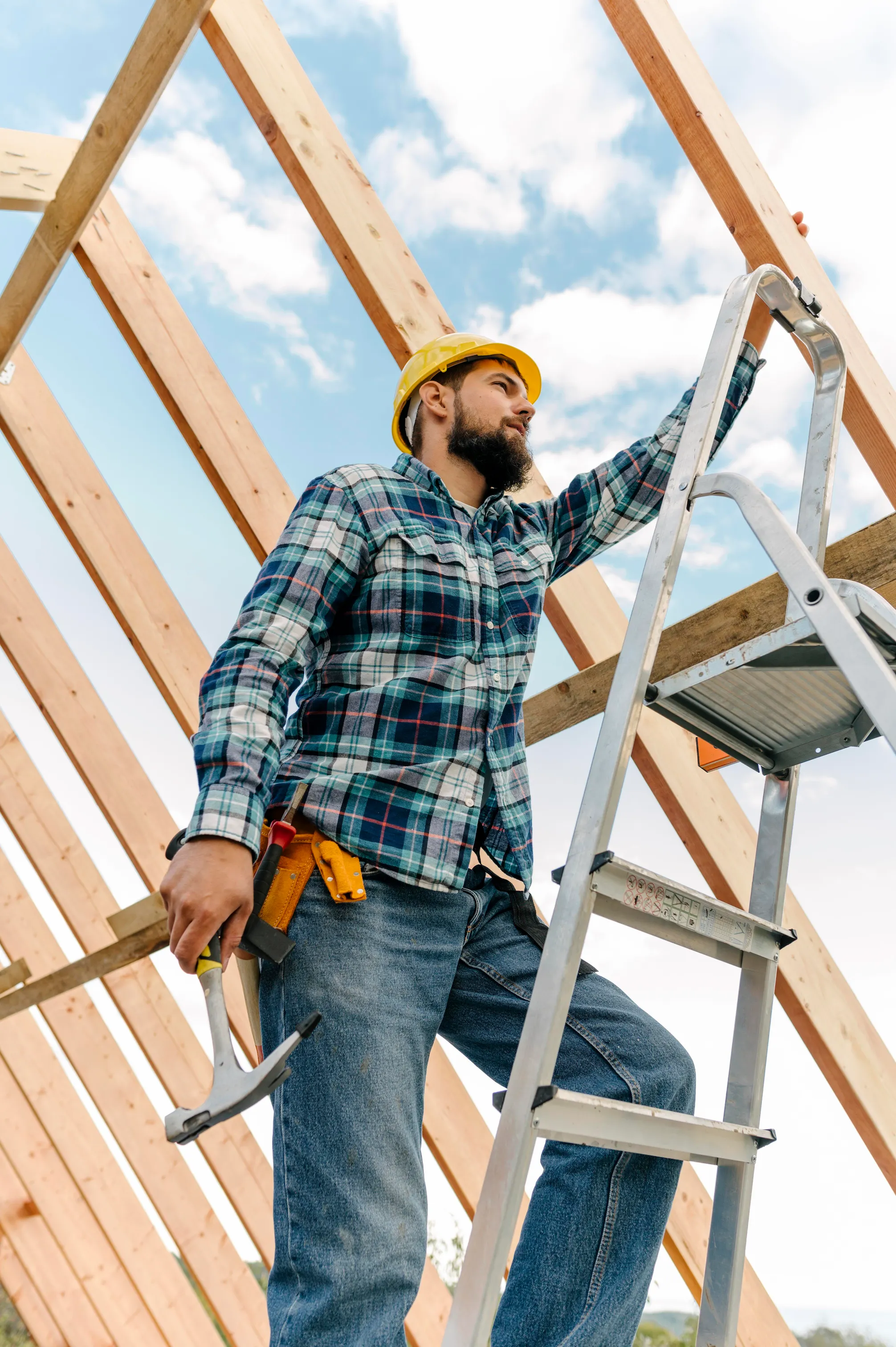 Construction worker wearing a yellow hard hat and plaid shirt standing on a ladder holding a hammer under a wooden frame roof.