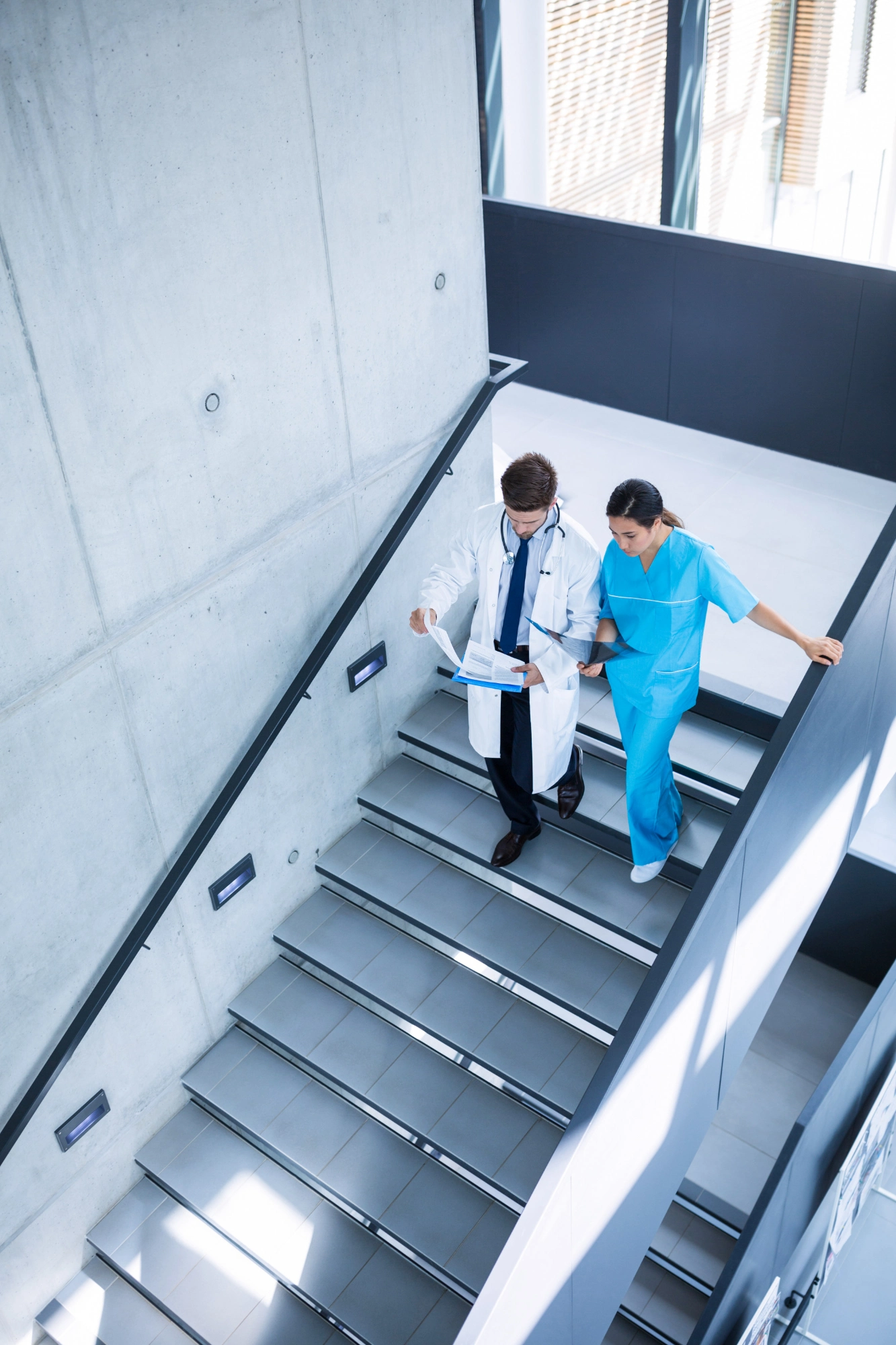 Two medical professionals, a doctor and a nurse, walking down a stairwell reviewing documents together.