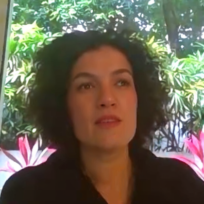 Woman with curly dark hair wearing a dark top sitting indoors with green plants and pink flowers in the background.