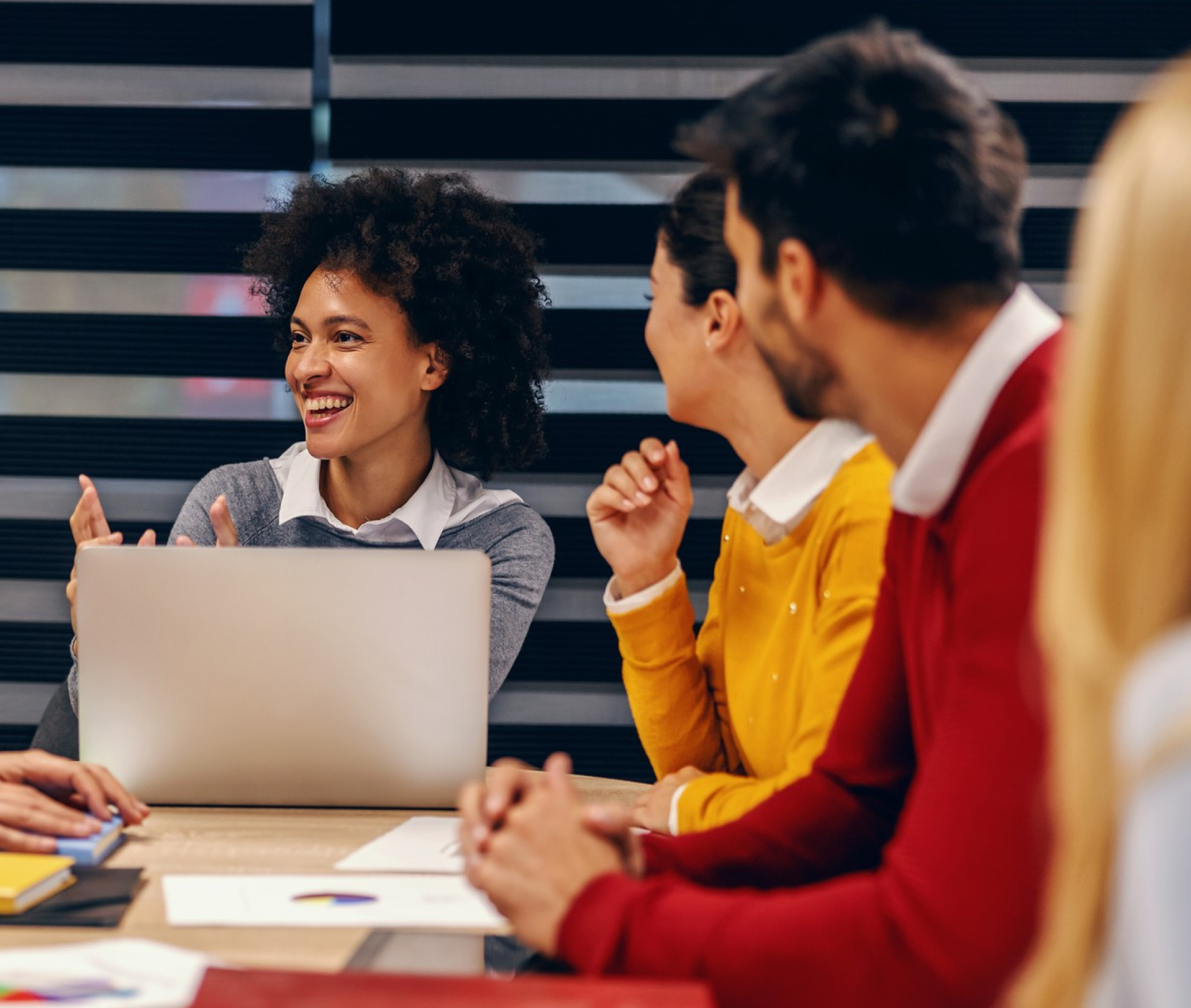 Smiling woman with curly hair gestures while talking to colleagues in a meeting with a laptop and documents on the table.