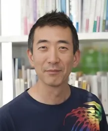 Smiling Asian man with short black hair wearing a dark blue shirt standing in front of a bookshelf.