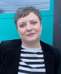 Smiling person with short grayish hair wearing a black jacket over a black and white striped shirt, sitting in front of a turquoise wall.