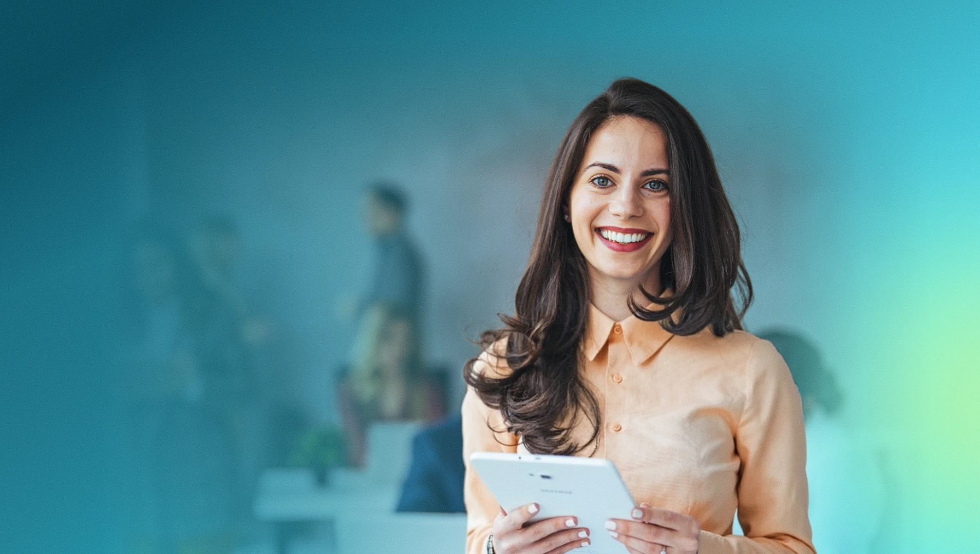 Smiling woman holding a tablet with blurred office colleagues in the background.