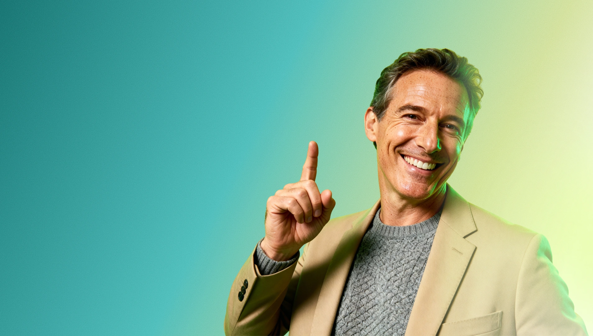 Smiling man in beige blazer and gray sweater pointing upward with his right index finger.