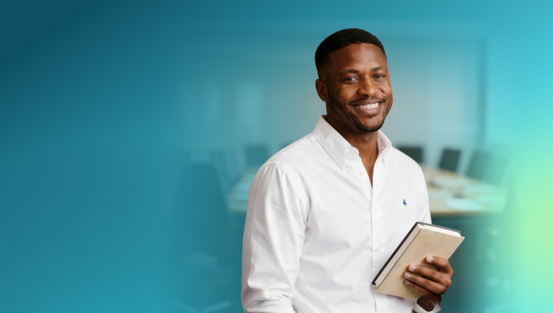Smiling man wearing a white button-up shirt holding a closed book in an office setting.
