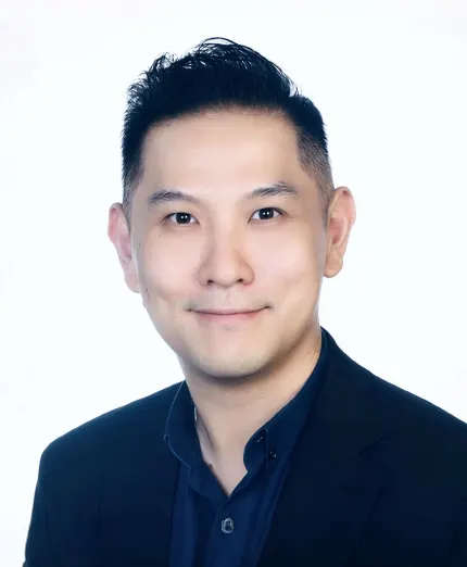 Portrait of a man with short black hair wearing a dark blazer and shirt, smiling slightly against a white background.