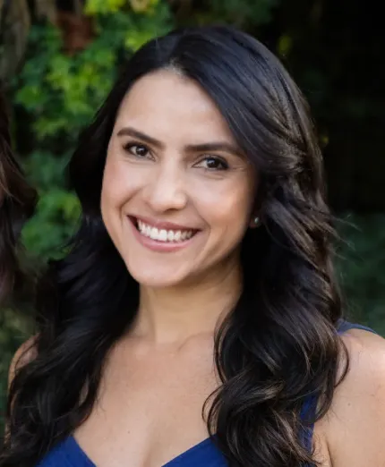 Smiling woman with long dark wavy hair wearing a blue sleeveless top, with greenery in the background.