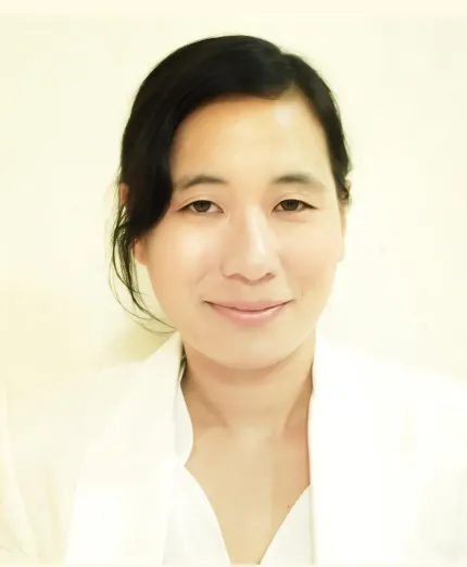 Portrait of a smiling woman with black hair wearing a white shirt and jacket against a light background.