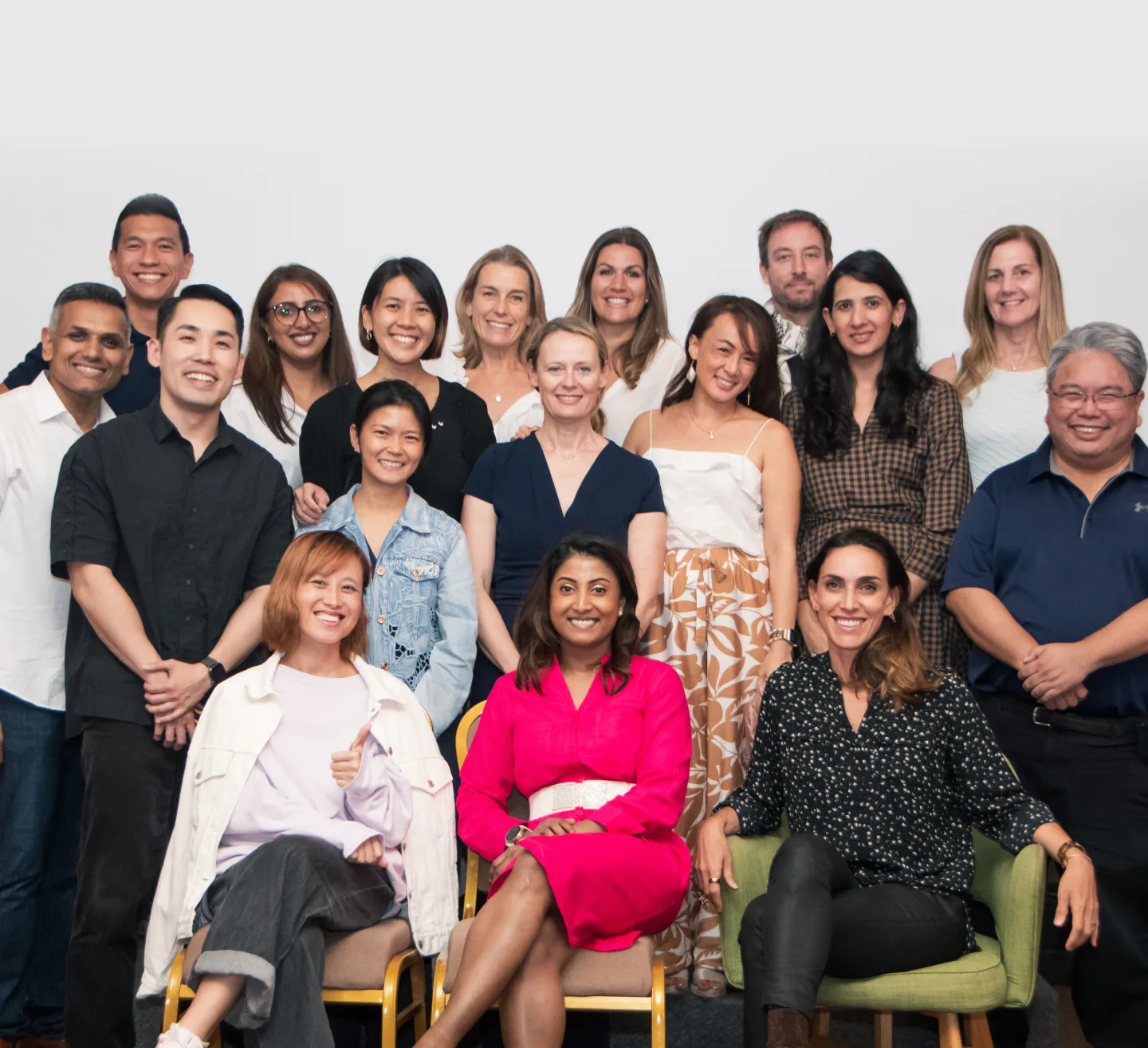 Group portrait of 18 diverse adults, some seated and others standing, all smiling against a plain light background.