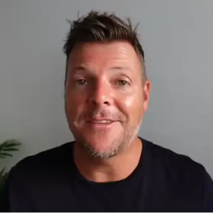 Smiling man with short brown hair and stubble wearing a black shirt against a gray background.