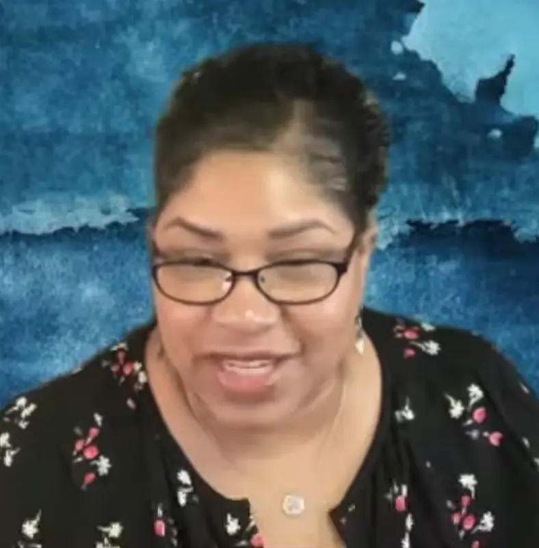 Woman with glasses and a black floral top smiling in front of a blue textured background.