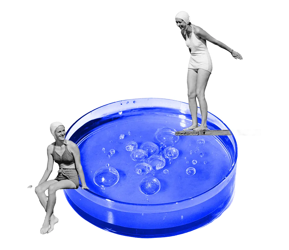 Black-and-white photo of two women in vintage swimsuits around a large blue petri dish with bubbles, one sitting on the edge and one standing on a diving board.