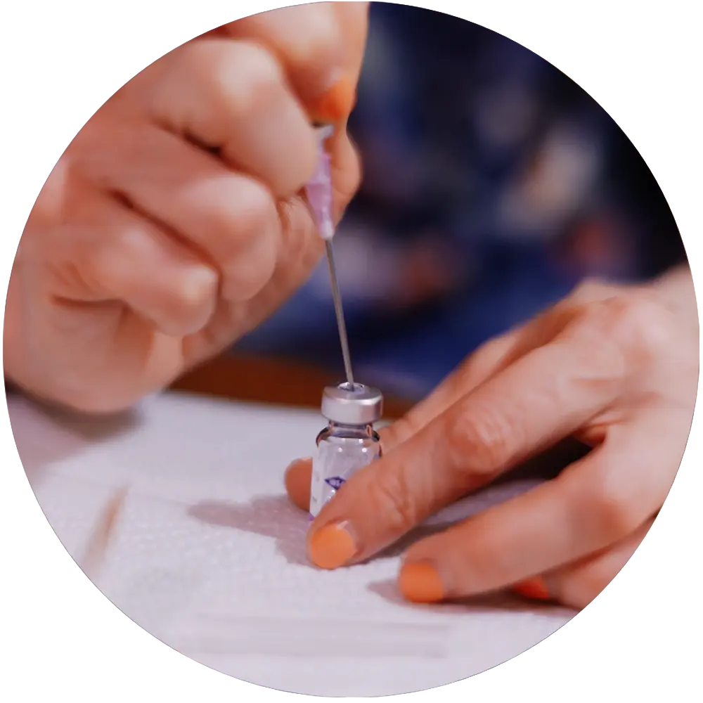 Hands with orange nail polish preparing a syringe by drawing liquid from a small vial on a white surface.