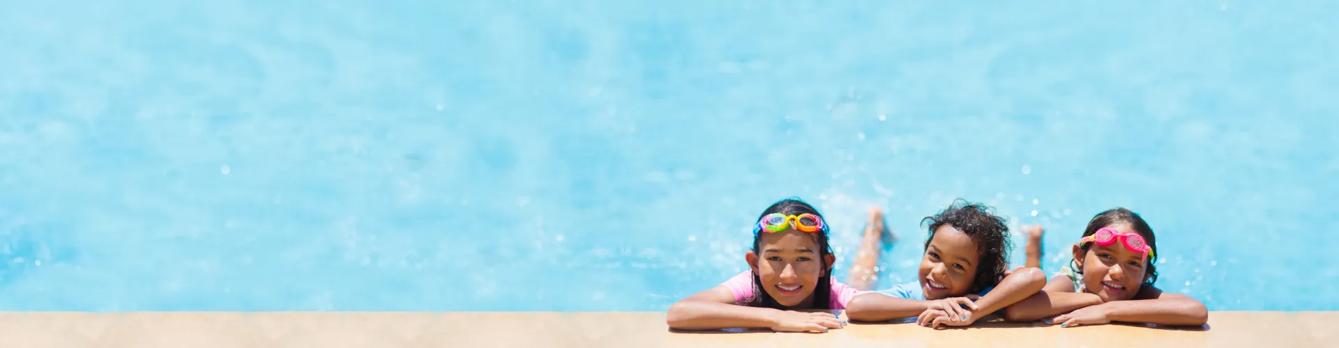Three children smiling and resting on the edge of a swimming pool with clear blue water.