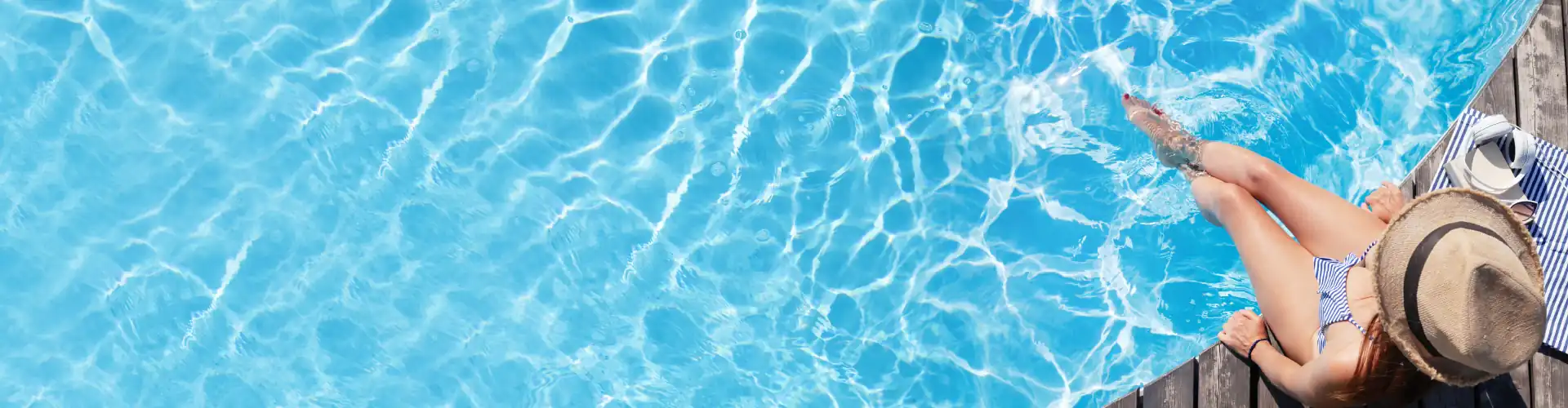 Woman in striped swimsuit and straw hat relaxing with feet in clear blue pool water beside wooden deck.