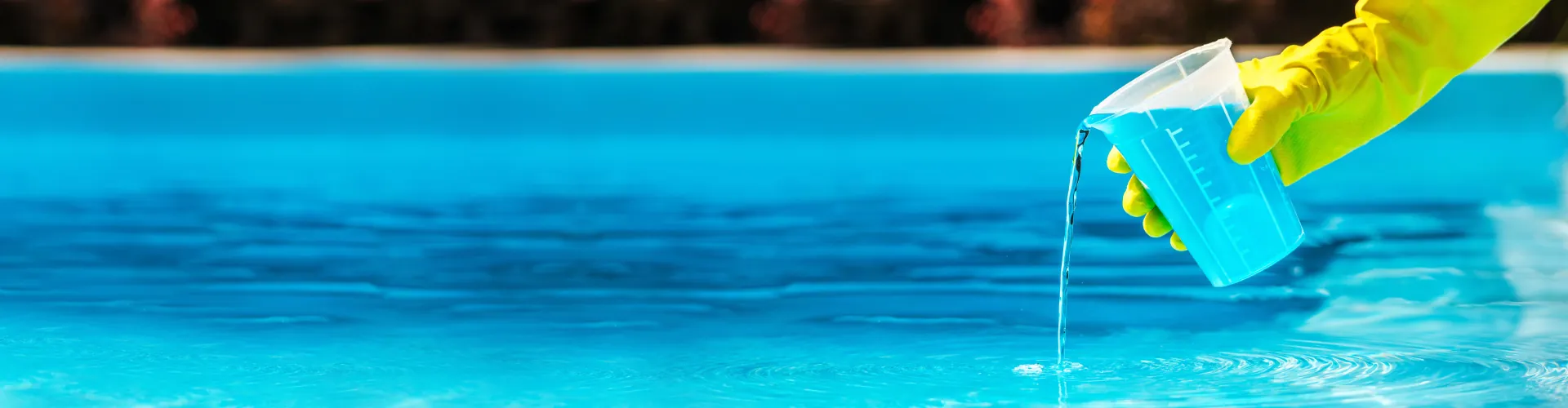 Hand wearing a yellow glove pouring blue liquid from a measuring cup into a swimming pool.