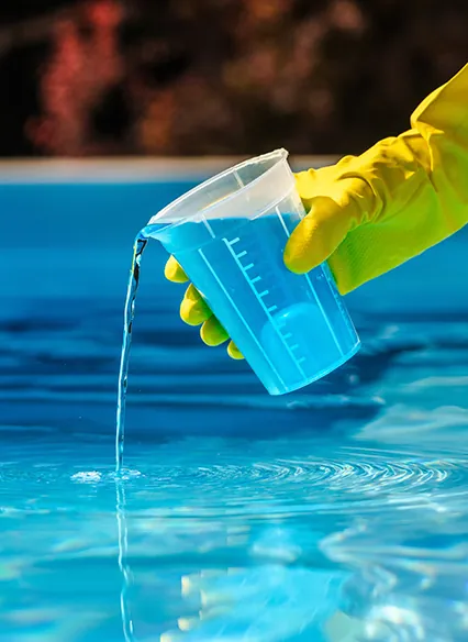Hand wearing yellow glove pouring blue liquid from a measuring cup into a pool.
