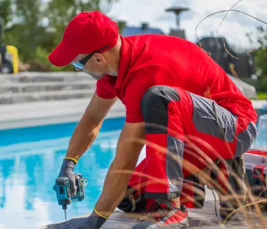 Man in red cap and shirt wearing gloves using a power drill by a pool.