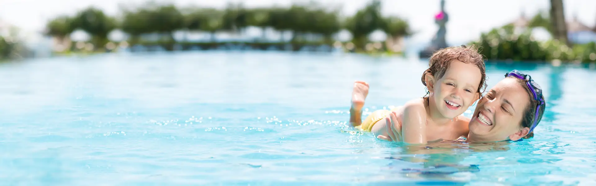 Smiling woman wearing swim goggles holding a happy child in a clear blue swimming pool.