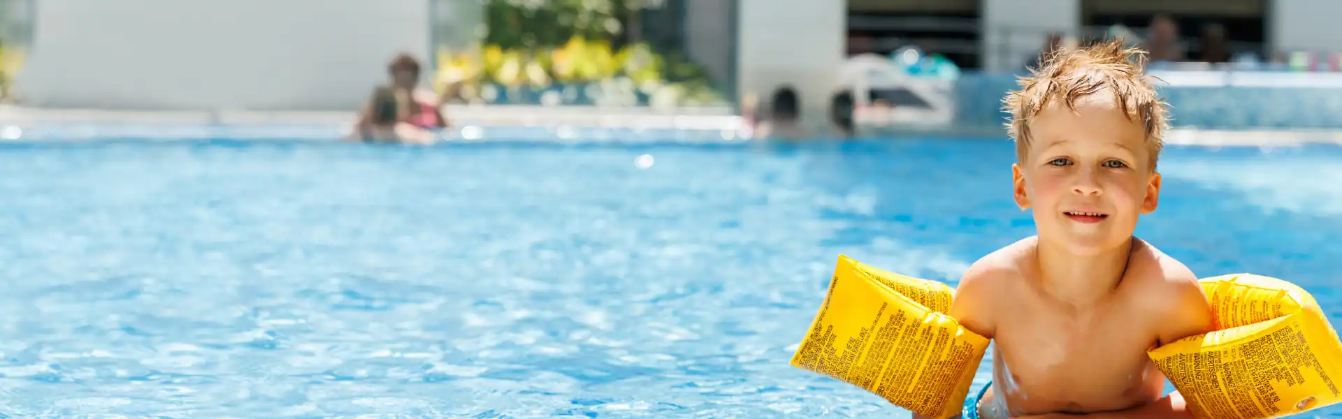 Young boy with yellow inflatable arm bands smiling in an outdoor swimming pool.
