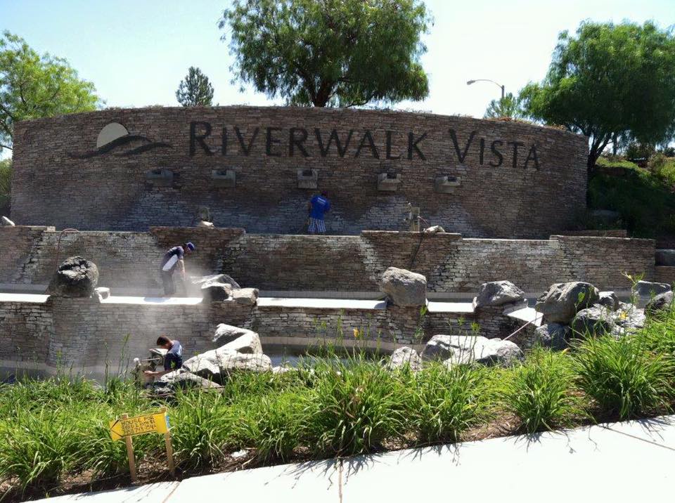 Riverwalk Vista entrance with stone wall, landscaping, and people working