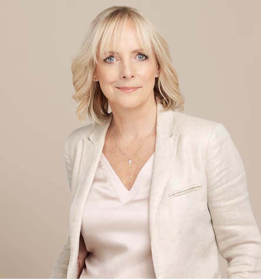 Blonde woman with blue eyes wearing a light-colored blazer and blouse against a beige background.