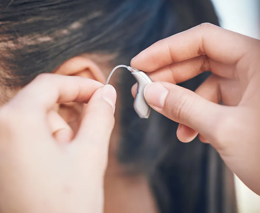 Hands holding and cleaning a small hearing aid device with a specialized tool on a work surface.