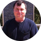 Smiling woman with short dark hair and glasses wearing a black blazer and white top sitting on a vintage metal bed frame.